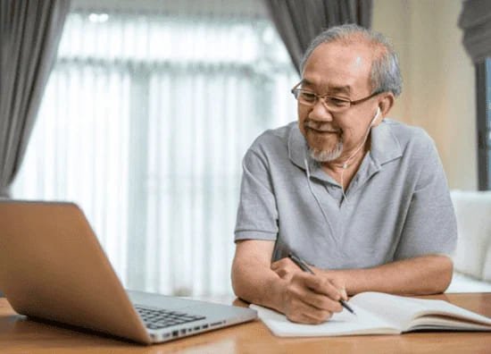 ノートを取りながらオンラインで英会話を学ぶシニア男性。